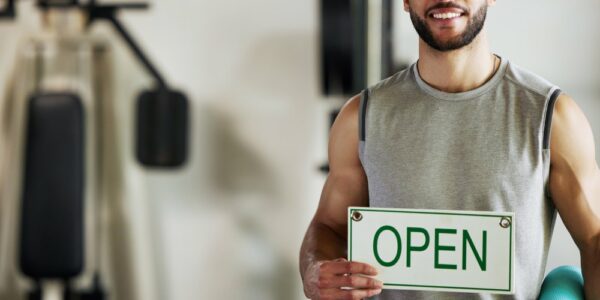 A man in a gray shirt holds a white open sign while holding a teal yoga mat and standing in a gym.