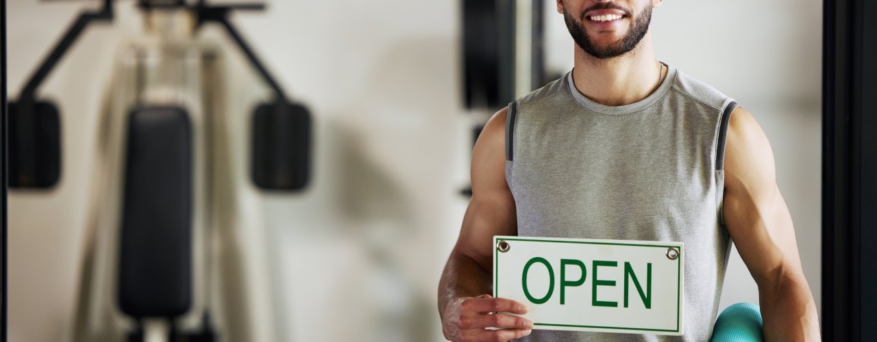 A man in a gray shirt holds a white open sign while holding a teal yoga mat and standing in a gym.