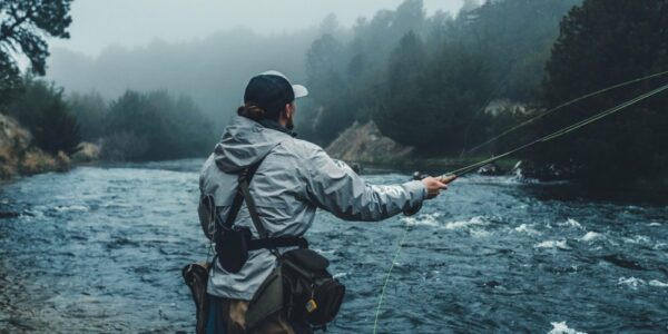 A man stands in a river, fishing. The water is moving downstream, and the weather is very misty and cloudy.