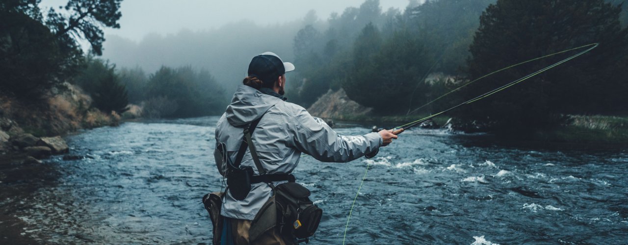 A man stands in a river, fishing. The water is moving downstream, and the weather is very misty and cloudy.