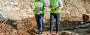 Two workers wearing hard hats and electric green vests walking along a dirt path with sticks and cut-up logs littering it.