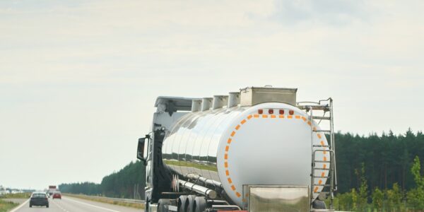 A tanker truck transporting liquid drives down a highway. Other cars are ahead of the truck with trees in the distance.