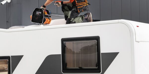A man in an orange shirt and hat holds a tool in one hand and tool bag in the other on the roof of an RV.