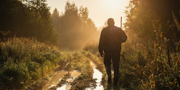 A hunter walking toward the sunset. He's next to a muddy dirt path with trees on both sides of him.