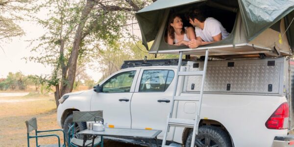 A young couple lies flat on their stomachs in a rooftop tent atop a white pickup truck at a desert campsite.