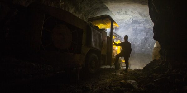 A person stands near a drilling machine as its light casts a glow across the dark underground mining tunnel.