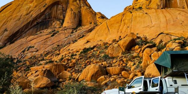 A white truck with a green rooftop tent parked in front of large rock formations with a blue sky in the background.