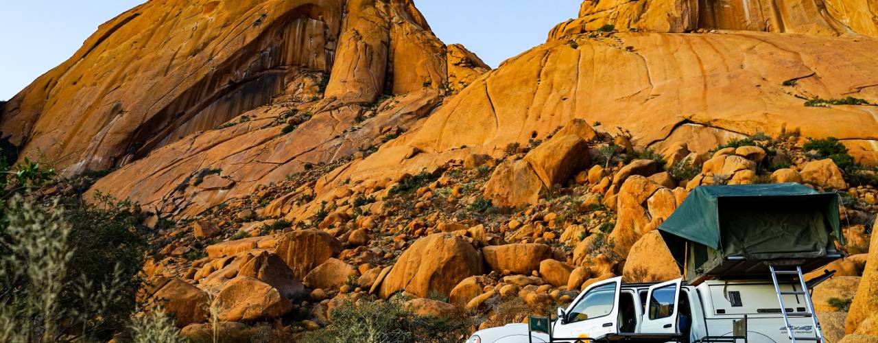 A white truck with a green rooftop tent parked in front of large rock formations with a blue sky in the background.