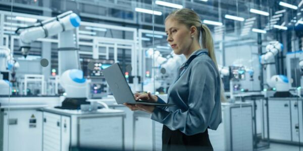 A woman standing on a factory floor holds a laptop in her hand. Robots assembling parts are behind her.
