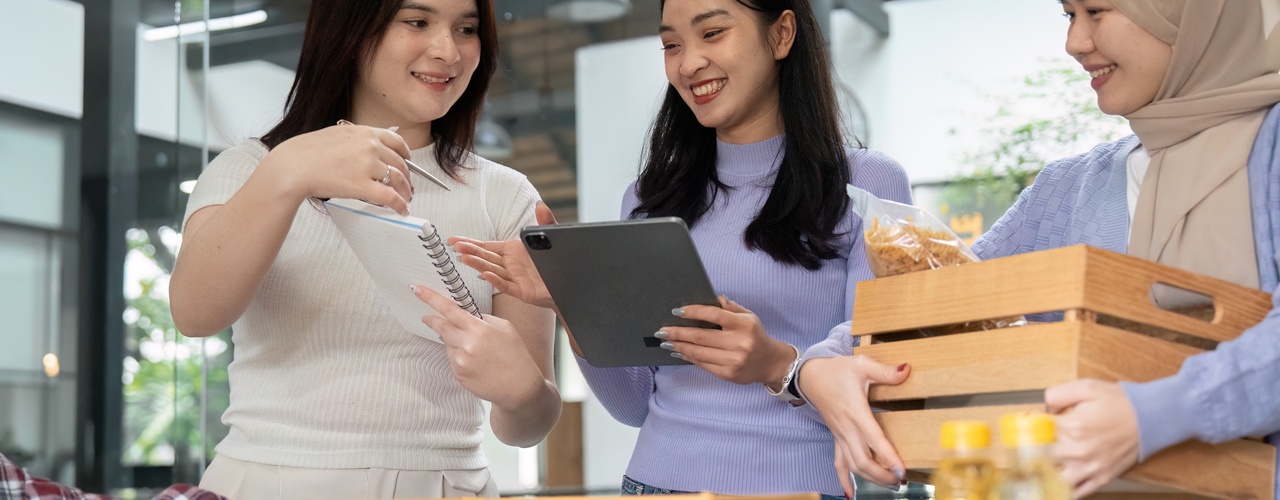 Three young, diverse women are laughing and talking while standing over a donation table with various donated items.