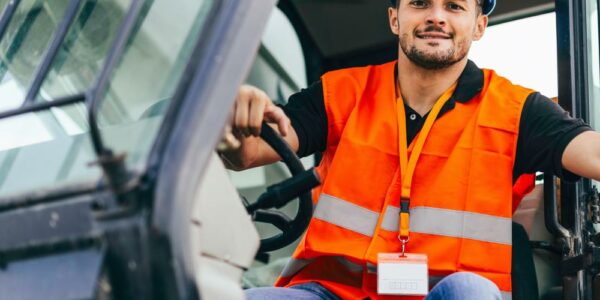 A construction worker in a neon orange safety vest smiles as he sits in the cab of a skid steer, his hand on the steering wheel.