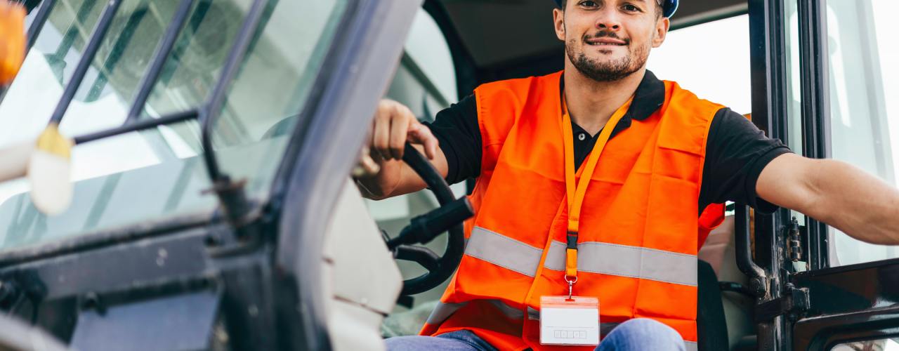 A construction worker in a neon orange safety vest smiles as he sits in the cab of a skid steer, his hand on the steering wheel.