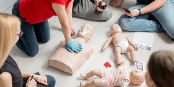A CPR instructor sits in a circle of students. She leans over as she showcases chest compressions on a manikin.
