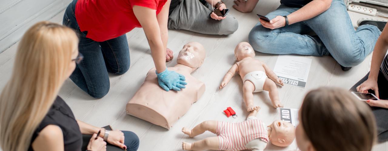 A CPR instructor sits in a circle of students. She leans over as she showcases chest compressions on a manikin.
