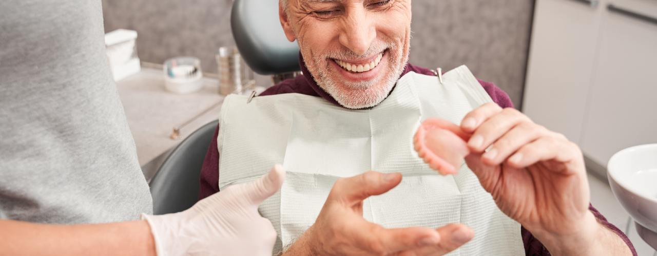 A senior man sits in a dentist chair as he holds a pair of dentures in his hand. He's smiling as he inspects them.