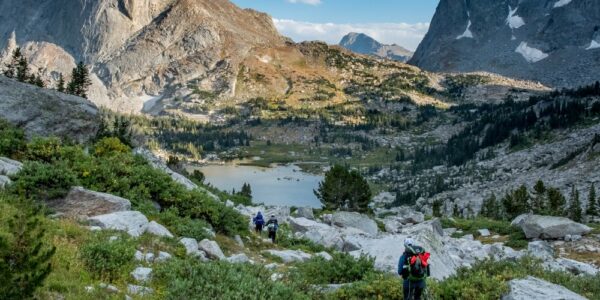 Three backpackers hike along the valley in the Cirque of the Towers in the Wind River Range, Wyoming. The sun sets in the distance.