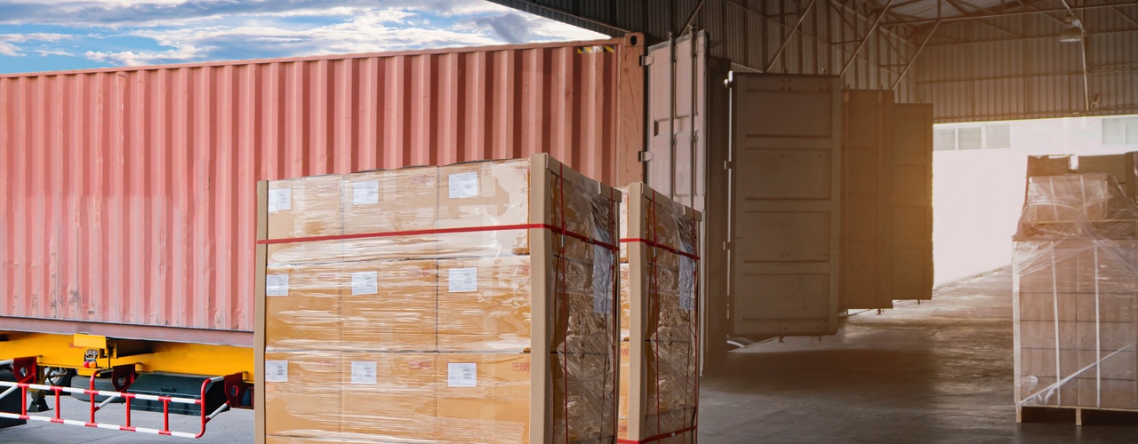 Two wooden pallets of goods, neatly stacked and wrapped, sit at a warehouse dock with freight trucks in the background.