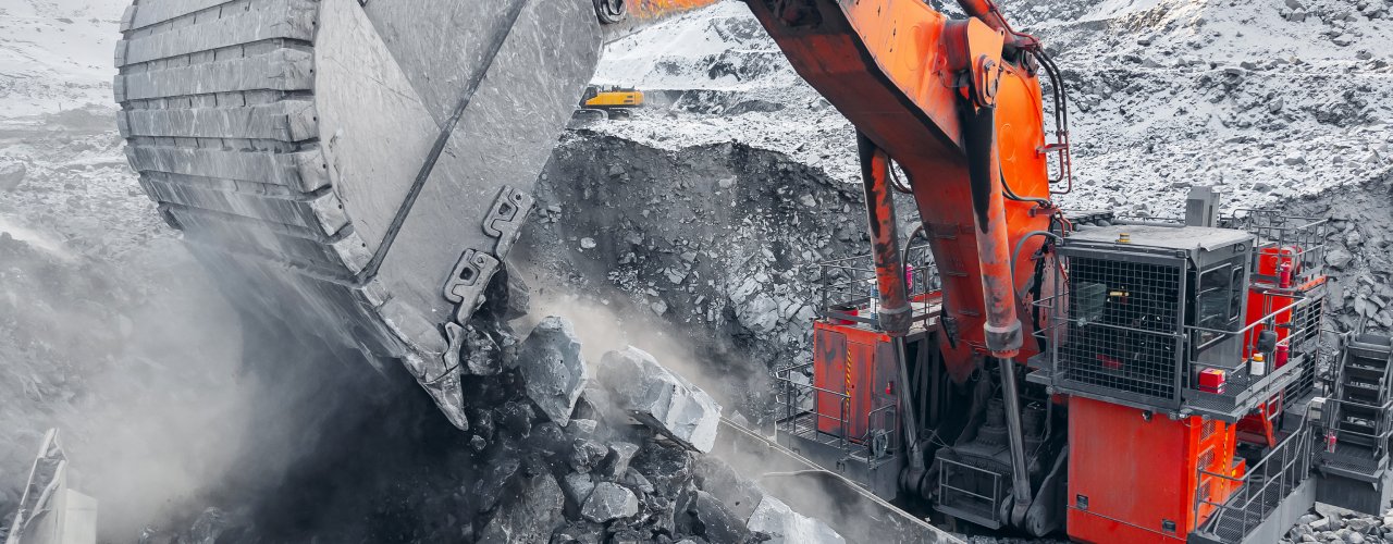 A red excavator dumping coal into the top of a dump truck. There is some snow on the piles of rocks in the background.