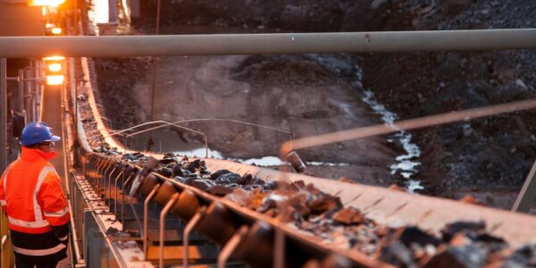 A mining worker standing next to a conveyor belt carrying ore rocks. The person is in a visibility jacket and a hard hat.