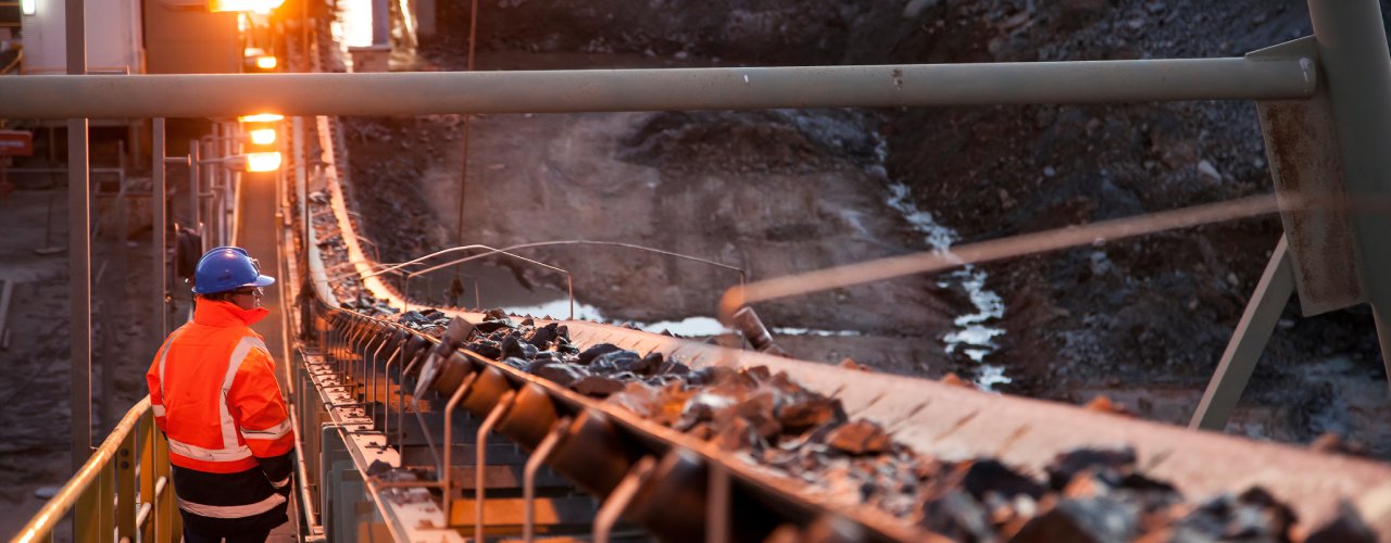 A mining worker standing next to a conveyor belt carrying ore rocks. The person is in a visibility jacket and a hard hat.