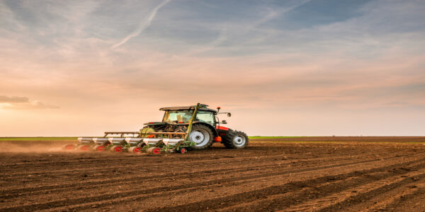 A person drives a red tractor drilling and seeding crops on an empty farm field underneath a cloudy sky.