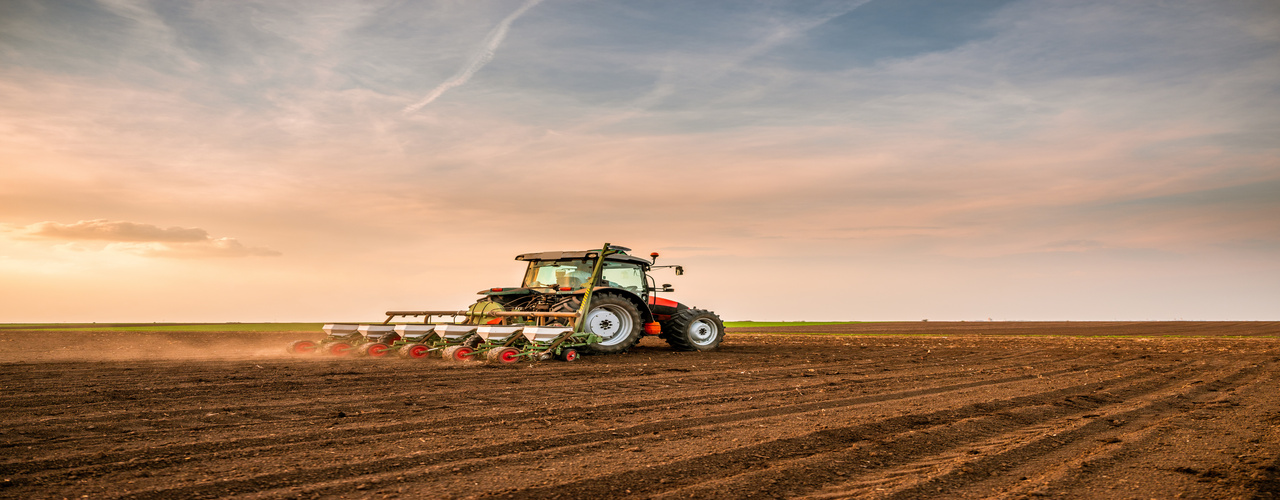 A person drives a red tractor drilling and seeding crops on an empty farm field underneath a cloudy sky.