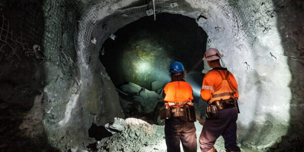 Two workers in orange vests stand inside an underground mine. They're using flashlights to look into the tunnel.