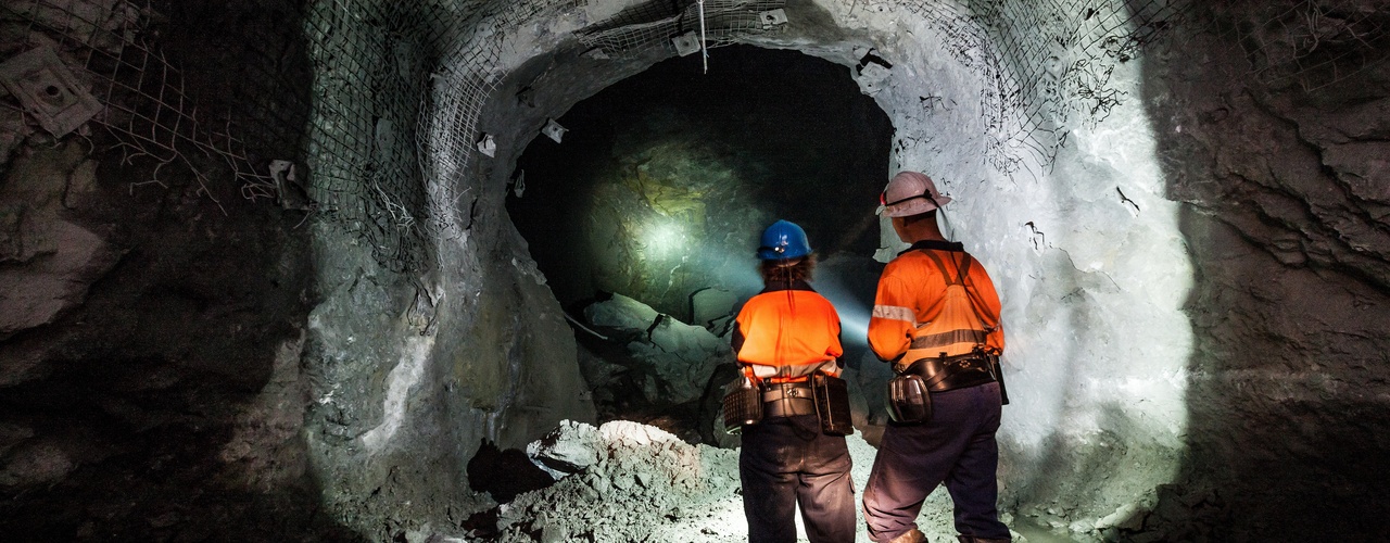 Two workers in orange vests stand inside an underground mine. They're using flashlights to look into the tunnel.