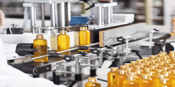A manufacturing assembly line for cosmetics. A worker wearing black gloves holds a gold bottle filled with a liquid.