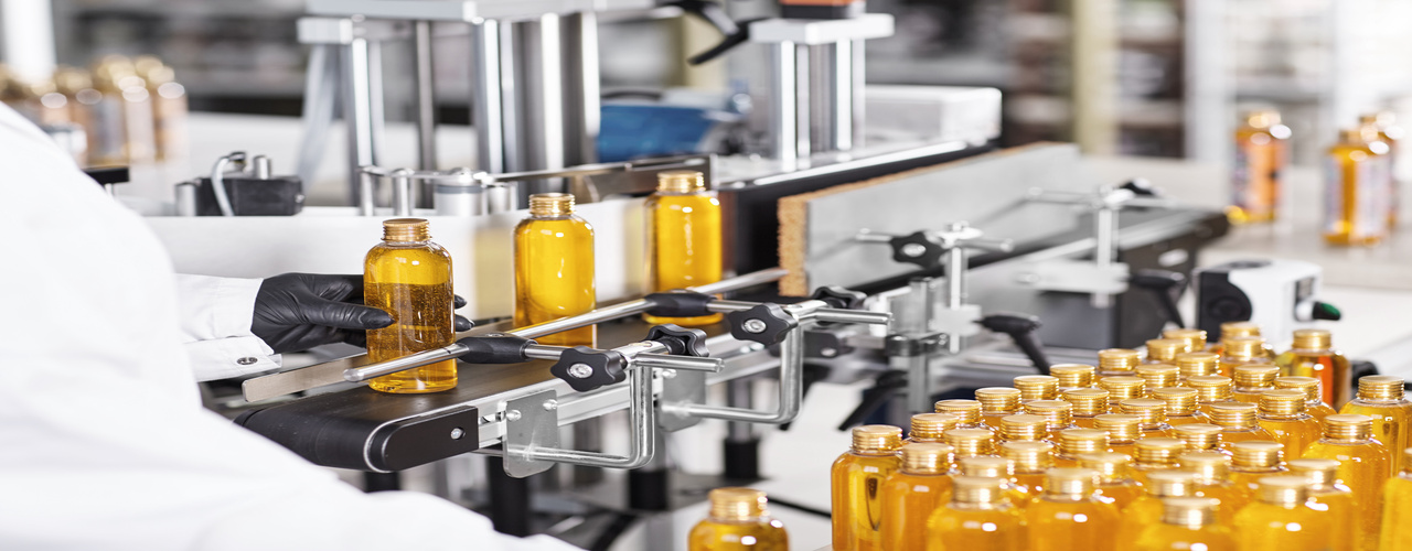 A manufacturing assembly line for cosmetics. A worker wearing black gloves holds a gold bottle filled with a liquid.