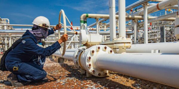 A worker in protective gear turning a pipeline valve. They are crouched down next to one part of a complex pipeline network.
