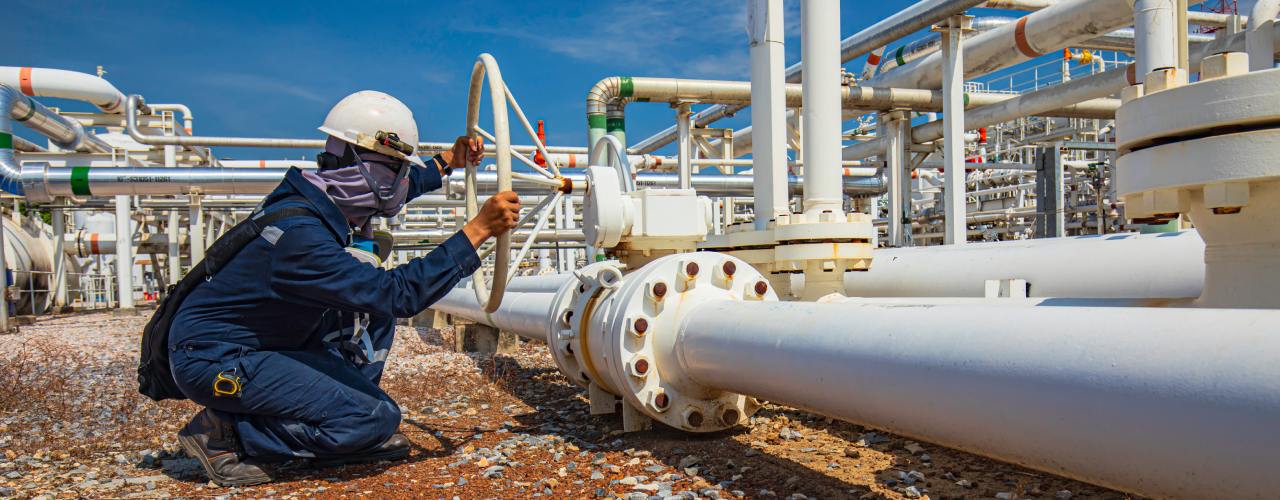 A worker in protective gear turning a pipeline valve. They are crouched down next to one part of a complex pipeline network.