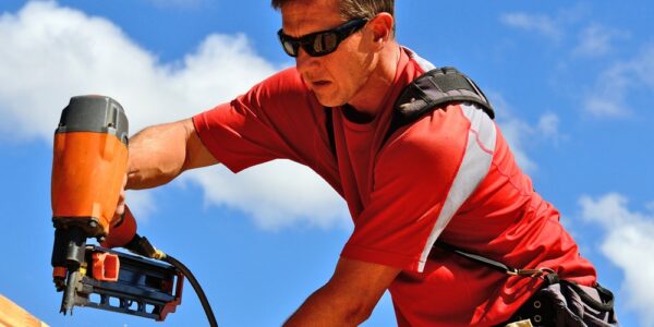 A man in a red T-shirt, khaki pants, and black sunglasses uses an orange nail gun to attach pieces of wood.