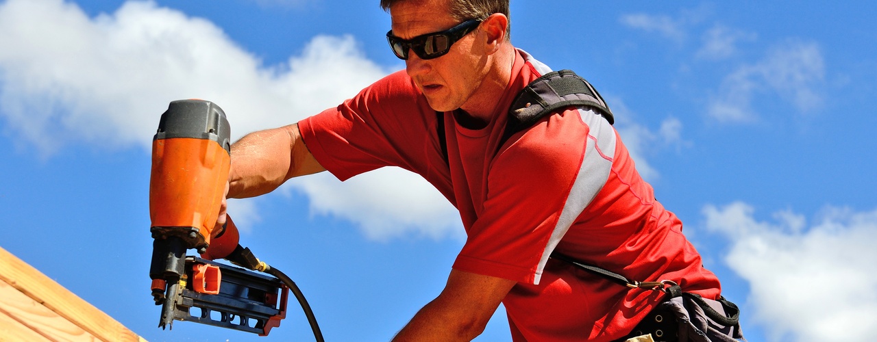 A man in a red T-shirt, khaki pants, and black sunglasses uses an orange nail gun to attach pieces of wood.