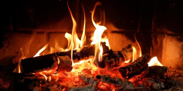 A close-up of a stone fireplace with burning wood logs and soot on the hearth. The flames cast a red glow.