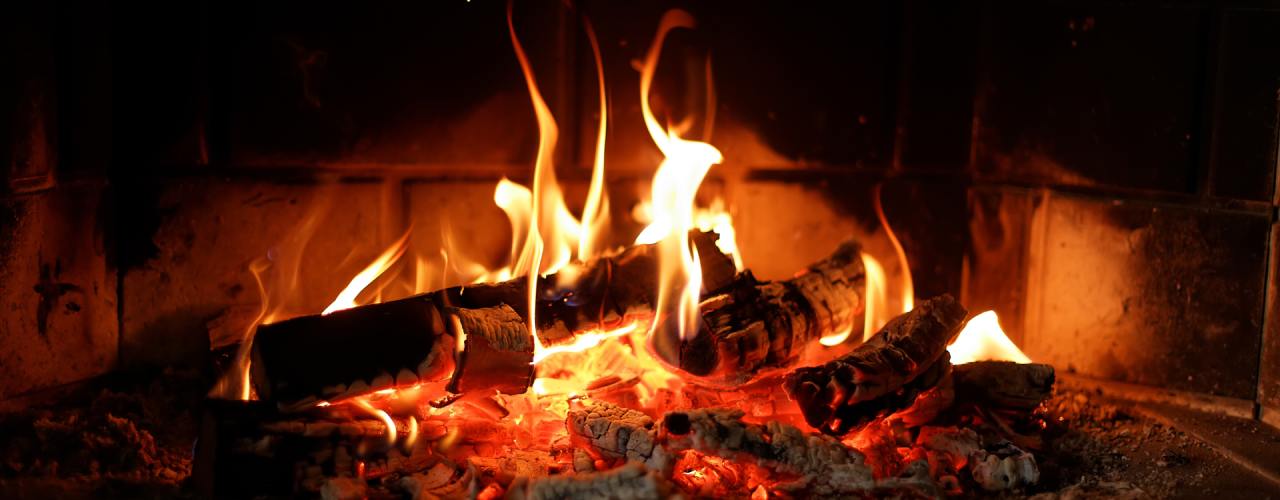 A close-up of a stone fireplace with burning wood logs and soot on the hearth. The flames cast a red glow.