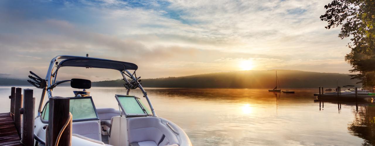 A boat with a white interior is moored to a wooden dock on a misty lake with the sun rising in the distance.