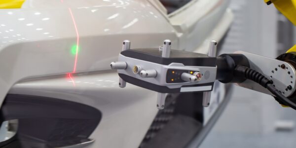 A yellow robotic arm scans a white car bumper with a red laser line and green dot during automated quality inspection.