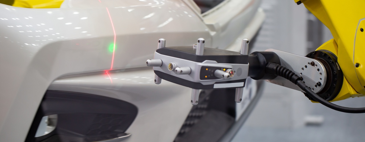 A yellow robotic arm scans a white car bumper with a red laser line and green dot during automated quality inspection.