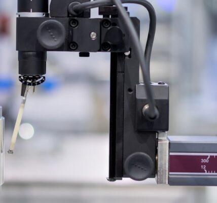 A coordinate measuring machine inspects a cylindrical metal part on a rotary table using a millimeter-marked probe arm.