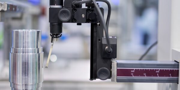 A coordinate measuring machine inspects a cylindrical metal part on a rotary table using a millimeter-marked probe arm.