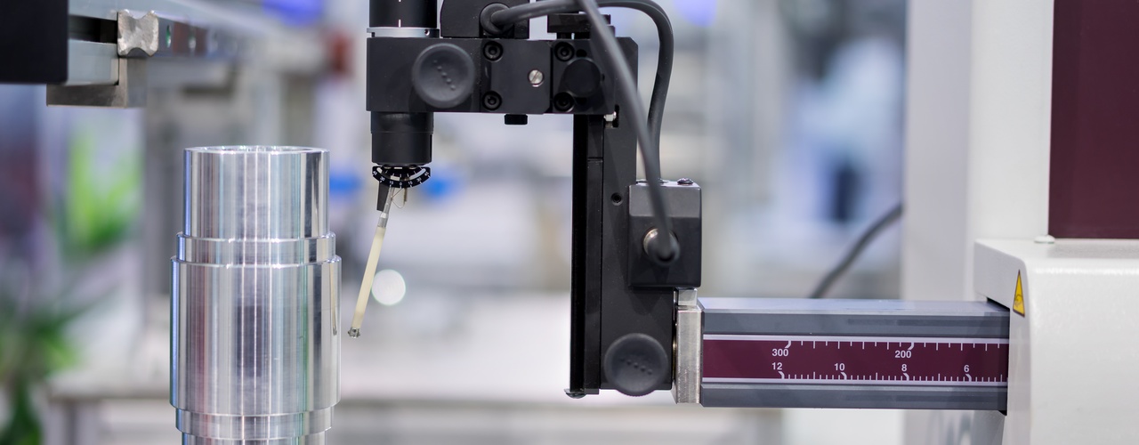 A coordinate measuring machine inspects a cylindrical metal part on a rotary table using a millimeter-marked probe arm.