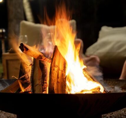 A close-up of an above-ground fire pit bowl with a burning stack of wood and someone sitting in a chair behind it.