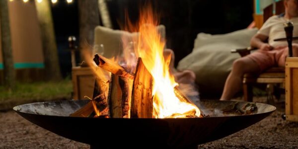 A close-up of an above-ground fire pit bowl with a burning stack of wood and someone sitting in a chair behind it.