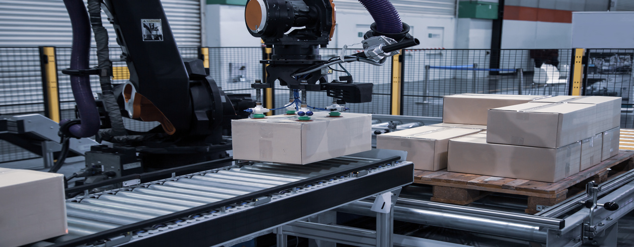 A black robot arm picking up cardboard boxes from a nearby pallet and placing them on a conveyor belt.