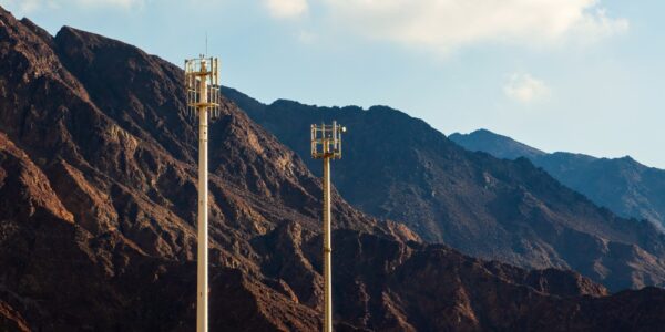Two tall cell towers with antennas stand before rugged mountains under a clear sky with scattered clouds.