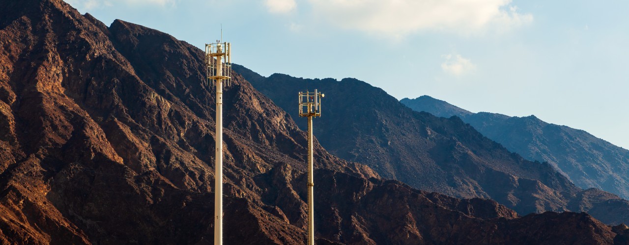 Two tall cell towers with antennas stand before rugged mountains under a clear sky with scattered clouds.