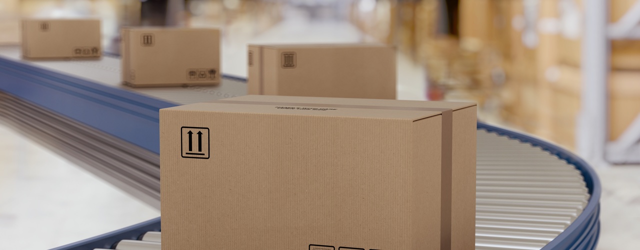 A close-up of a taped cardboard box on a roller conveyor belt in a shipping facility with more boxes behind it.