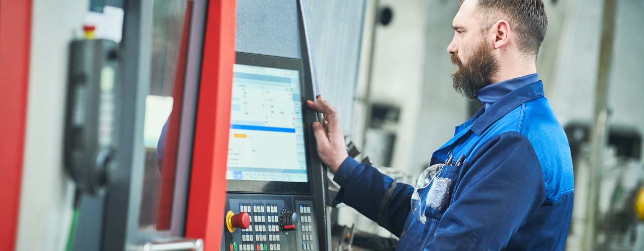 A man with a brown beard and short hair is standing next to an industrial machine. He is looking at a monitor.