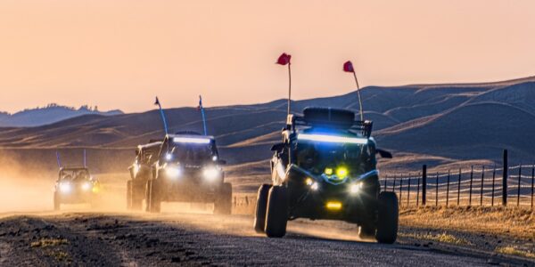 A line of four UTVs driving on a dirt road. There are large dunes of sand behind them in the distance.